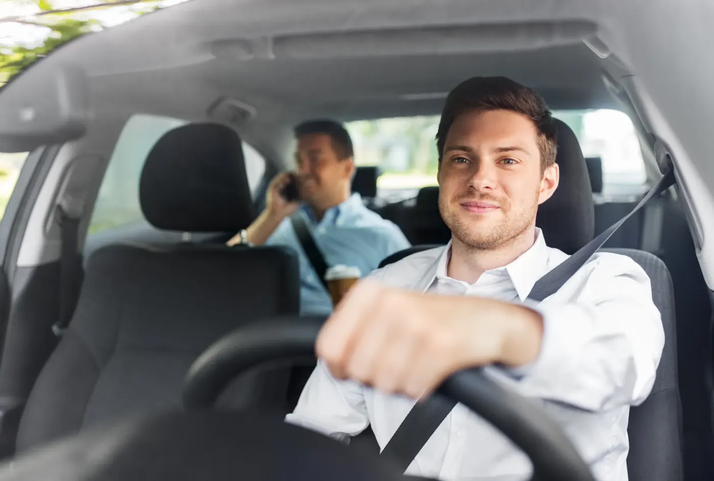 Two occupants seated in a car, both wearing seatbelts, viewed through the windscreen with the driver holding the steering wheel.