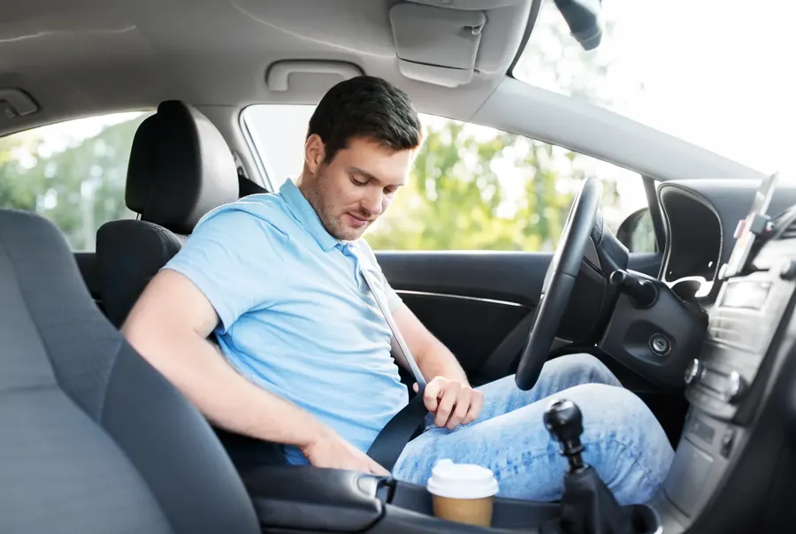 A man seated in the driver's seat of a car, drawing the seatbelt across his chest towards the buckle.
