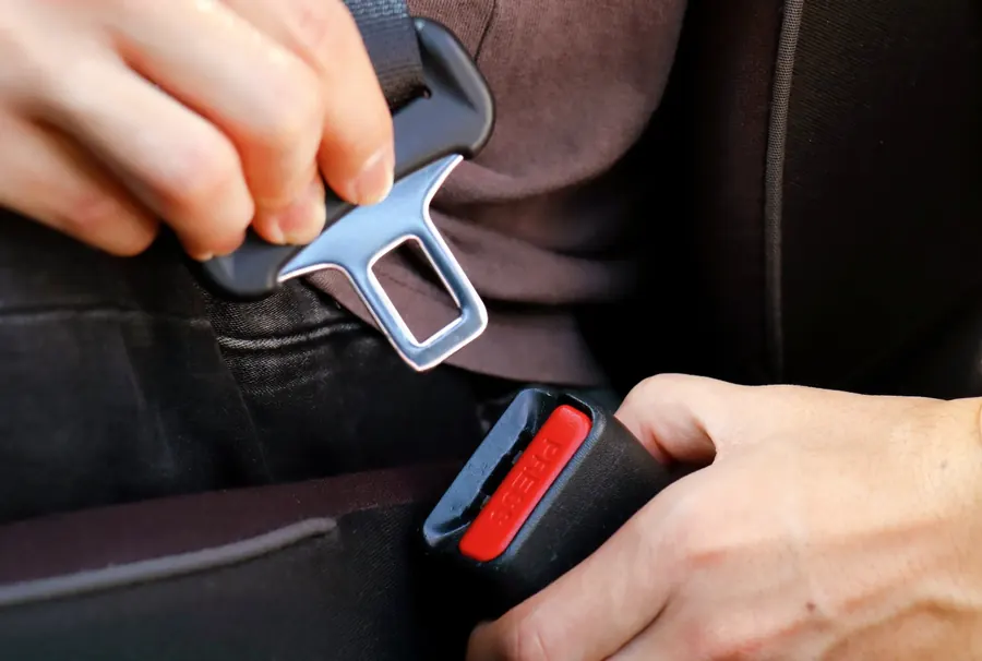 Two hands guiding a metal seatbelt latch plate into a black buckle receiver marked PRESS.