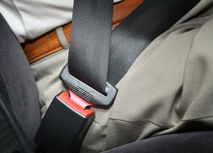 A fastened seatbelt across a seated person's lap, with the black latch plate locked into the buckle receiver beside the seat.