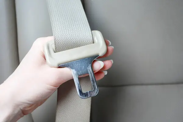 A hand holding a seatbelt latch plate with the webbing running through it, against a grey vehicle seat.