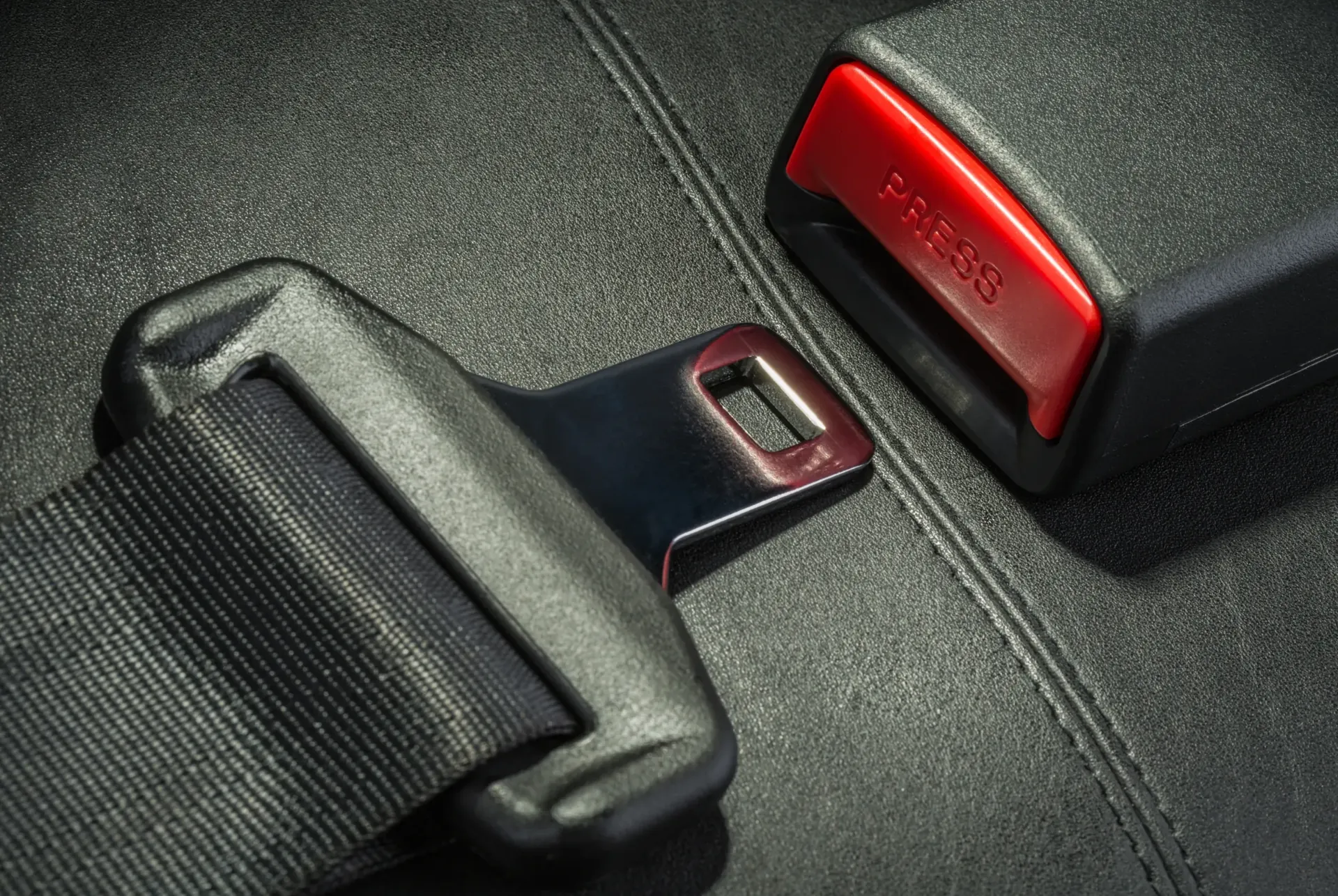 A vehicle seatbelt latch plate resting on dark leather upholstery beside its buckle receiver, with the red release button clearly marked PRESS.