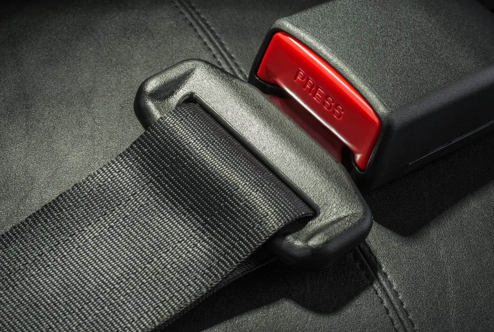 A seatbelt assembly photographed close up on dark upholstery, showing woven webbing entering a moulded latch plate next to the buckle receiver.