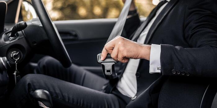 A person in a dark suit seated in a car, holding the seatbelt latch plate in one hand before fastening it.
